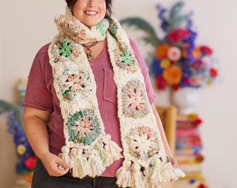 May include: A cream-colored, crocheted scarf with colorful floral squares and fringe. The scarf is worn over a pink striped shirt and dark gray jeans. The scarf's design includes pastel pink, green, and blue accents.