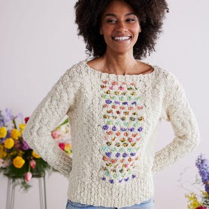 May include: Cream-colored knit sweater with a textured pattern and a scoop neckline. The front features a vertical floral embroidery in various colors. The sleeves are long, and the sweater is paired with blue jeans. The overall style is casual and comfortable.