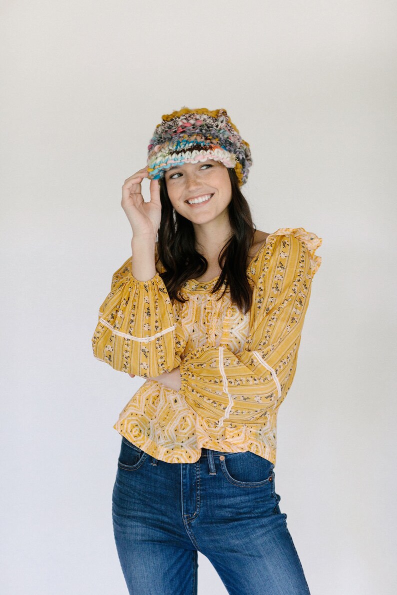 May include: A woman wearing a yellow floral blouse with white trim, blue jeans, and a colorful crocheted hat.