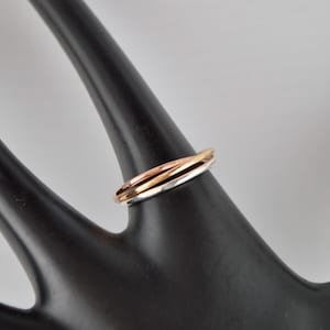 May include: A tri-color ring with intertwined bands of gold, rose gold, and silver. The ring is displayed on a black, stylized hand, showcasing its design and metallic hues. The ring's bands are smooth and polished.