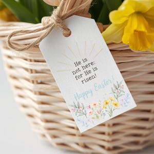 May include: A woven basket with a tag tied with twine. The tag reads "He is not here: for He is risen! Happy Easter!" with floral accents. Yellow daffodils and pink tulips are visible.