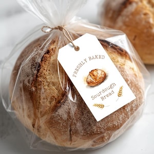 Könnte beinhalten: Ein rundes Sauerteigbrot in einer transparenten Plastiktüte, mit Bindfaden gebunden. Ein weißes Etikett mit der Aufschrift "FRISCH GEBACKEN" und "Sauerteigbrot" mit einer Brot- und Weizenillustration.