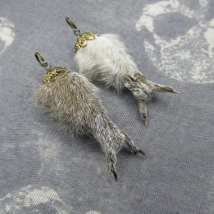 May include: Two rabbit's foot charms with brass-coloured metal accents. One charm is grey, and the other is white. The charms have a loop at the top for hanging. The rabbit feet are attached to a decorative brass-coloured metal piece.