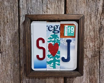 May include: A square wall art piece featuring three vertical segments of license plates framed in dark wood. The left segment shows a red letter S, the center features a green pine tree and a red heart, and the right shows a blue letter J with a Utah 19 sticker.
