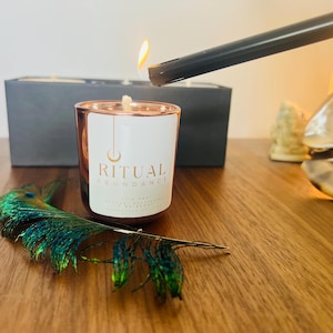May include: A lit candle in a rose gold glass with the text "RITUAL ABUNDANCE" on a white label. A peacock feather and a lighter are next to the candle. A box of candles is in the background.
