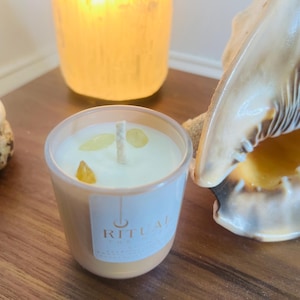 May include: A small, lit candle in a clear glass container with a white label that reads "RITUAL". The candle has a white wax with yellow crystals. A lit candle and a seashell are in the background.