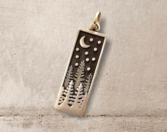 Puede incluir: Un colgante de plata con un diseño rectangular que presenta una luna creciente, estrellas y una silueta de pinos.