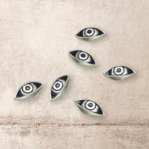 May include: Six silver eye charms with black detailing. The charms are shaped like an eye with a black pupil and iris.