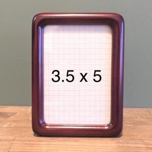 May include: A rectangular wooden picture frame with rounded corners. The frame is a dark reddish-brown colour. The dimensions are 8.9 x 12.7 cm. The interior has a white grid pattern.