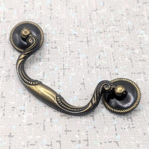 以下が含まれることがあります： カーブしたハンドルと装飾的な円形の端部を備えたアンティーク真鍮の引き出しプル。端部は暗く、中央に金色のノブとロープのようなボーダーが付いています。ハンドルは明るい真鍮色です。