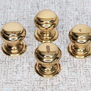 May include: Four gold-toned cabinet knobs with a rounded top and a slightly raised rim. The knobs have a smooth, polished finish.