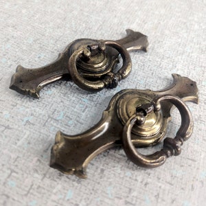 May include: Two antique brass drawer pulls with ring handles. The pulls have a decorative backplate with a curved design. The ring handles are attached to a circular base. The brass has a slightly aged appearance.