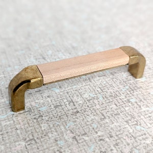 以下が含まれることがあります： 明るい木製のトップが付いた真鍮製のキャビネットプル。プルは丸みを帯びた長方形の形をしています。