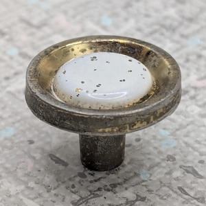 May include: A vintage cabinet knob with a white, speckled center and a gold-toned rim. The knob has a dark, aged metal base and stem, suitable for furniture restoration or adding a touch of antique charm to any project.