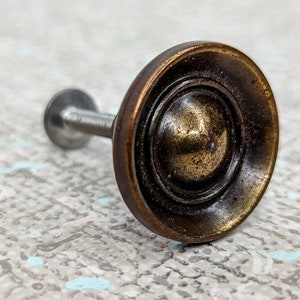 May include: A round, brass cabinet knob with a raised, concentric circle design. The knob has a silver screw.