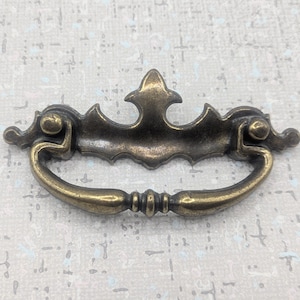 May include: Antique brass drawer pull with a decorative design. The pull features a curved handle and ornate detailing, including a central finial and decorative end pieces. The hardware is made of metal and has a vintage aesthetic.