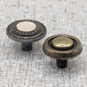 May include: Two decorative cabinet knobs. One knob has a white speckled centre with a dark grey border and a dark grey stem. The other knob has a brass-coloured centre and border with a dark grey stem.