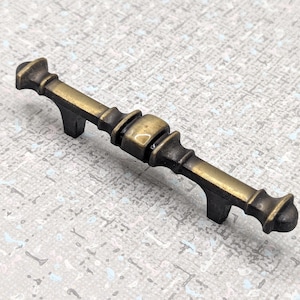 May include: Antique brass finish cabinet pull with a decorative design. The hardware features a central barrel shape and two curved feet. The pull is approximately 13 cm long.