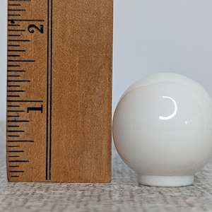May include: A white, spherical object with a glossy finish, approximately 1.5 inches in diameter, sits next to a wooden ruler. The ruler shows inch measurements. The object has a small, circular base and reflects light.