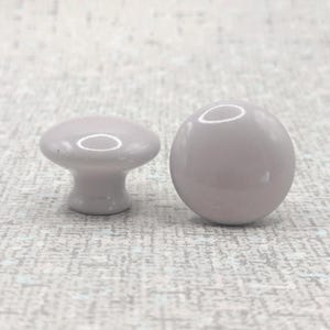 May include: Two light grey ceramic cabinet knobs. One is mushroom-shaped with a flat top and a short stem. The other is spherical. Both have a glossy finish against a textured background.