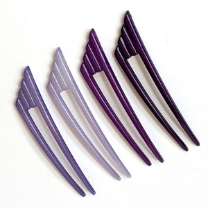 May include: Four purple hair sticks in varying shades, from light lavender to deep violet. Each stick features a fan-shaped top with vertical lines and a long, slender prong. The hair accessories are arranged diagonally against a white background.