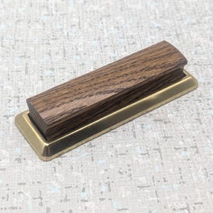 May include: A rectangular wooden desk nameplate with a brass base. The wood is a dark brown colour with visible grain. The brass base is a shiny gold colour. The nameplate is a simple, elegant design.