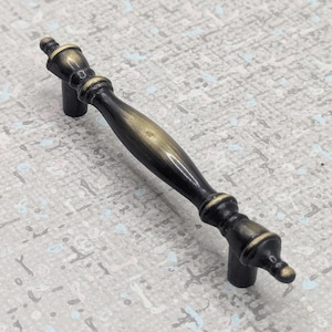 May include: A cabinet pull with a dark bronze finish and a gold-toned central section. The handle has a traditional design with detailed ends and a curved centre, measuring approximately 13 cm in length.