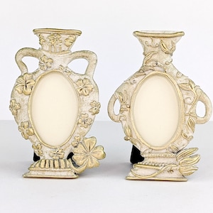 May include: Two white ceramic picture frames shaped like vases with gold accents and floral designs. The frames have oval openings for inserting photos.