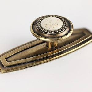 Peut inclure: Un bouton de porte en laiton avec un centre en céramique blanche et une bordure perlée décorative. Le bouton est fixé à une plaque rectangulaire en laiton.