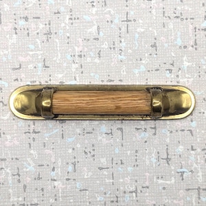 May include: A brass cabinet pull with a natural wood handle. The pull has a rectangular shape with rounded ends and a central wooden dowel. The brass has a slightly aged appearance, and the wood grain is visible.