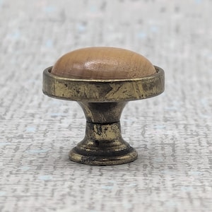 May include: A small, antique-style pin cushion with a light brown, rounded top. The base is made of aged brass-coloured metal, featuring a decorative, pedestal-like design. The pin cushion is a sewing accessory.