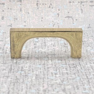 May include: A brass-coloured cabinet pull with an arched design. The rectangular top connects to two rounded legs, creating a bridge-like appearance. The hardware has a slightly aged, vintage look, suitable for furniture restoration or new builds.