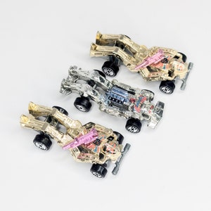 May include: Three toy cars in gold and silver metallic finishes with black wheels. The cars have intricate designs and are arranged on a white surface. One car has a pink accent.