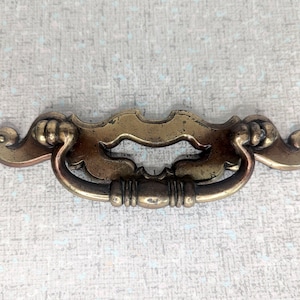 May include: A vintage brass drawer pull with a decorative, ornate design. The pull has a curved, arched shape with a central handle.
