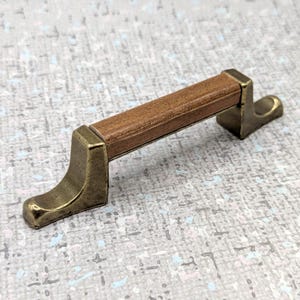 May include: A cabinet pull with a rectangular, wood-grain center and antique brass-colored metal brackets. The handle is a warm brown, and the metal has a slightly curved design.