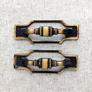 May include: Two antique brass drawer pulls with a black finish and a decorative design. The pulls have a rectangular shape with rounded corners and a raised center section.