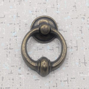 May include: Antique brass drawer pull with a ring handle. The hardware features a circular backplate and a rounded ring for easy gripping. The finish has a vintage look, suitable for cabinets and furniture.