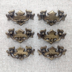 Peut inclure: Six poignées de tiroir en laiton antique avec un motif de feuille décoratif. Chaque poignée a une poignée incurvée et deux vis décoratives.