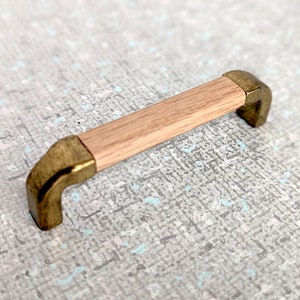 以下が含まれることがあります： 真鍮のアクセントが付いた木製のキャビネットプル。プルは丸みを帯びた角を持つ長方形で、明るい木製の仕上げになっています。真鍮のアクセントはプル両端にあり、わずかに古びた外観をしています。