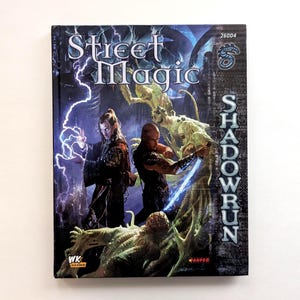 May include: A hardcover book titled "Street Magic" with the word "Shadowrun" on the side. The cover features fantasy art of characters with glowing energy and creatures in shades of green and blue.