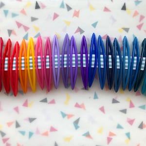 May include: A set of 28 colorful plastic crochet hooks arranged in a rainbow order. The hooks are all the same size and shape, and they are held together by a white and green striped ribbon.