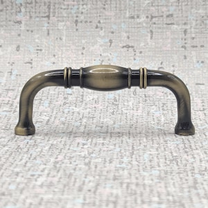 以下が含まれることがあります： アンティーク真鍮のキャビネットプル。曲線的なハンドルデザイン。中央部分は湾曲した端よりもわずかに幅が広く、両側に装飾的なリングが付いています。ヴィンテージ感のある仕上がりです。