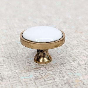 Peut inclure: Un bouton de meuble avec un dessus rond en céramique blanche et une base en métal doré. Le bouton a un design classique, adapté aux tiroirs ou aux armoires. La base a un aspect texturé et antique.