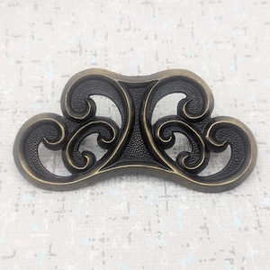 以下が含まれることがあります： 暗いブロンズ仕上げの、装飾的なアンティークスタイルの引き出しプル。装飾的なハードウェアは、渦巻くスクロールとテクスチャード加工された中央部の対称的なデザインが特徴で、家具にヴィンテージ感を加えるのに最適です。