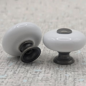 May include: Two white ceramic cabinet knobs with a dark, antique-style metal base. The knobs have a rounded, oval shape and a small, dark metal accent on top. These decorative knobs are ideal for furniture upgrades.