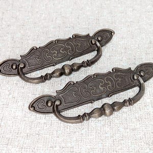 May include: A pair of antique bronze drawer pulls, featuring intricate scrollwork and a curved handle. The decorative backplates and detailed handles give a vintage feel. These cabinet pulls are suitable for furniture restoration, with each pull measuring approximately 15 cm in length.
