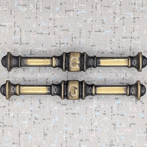 May include: A pair of antique cabinet pulls. Each pull features a dark bronze finish with a gold-toned central section and end caps. The rectangular pulls have a decorative design and are about 13 cm in length.