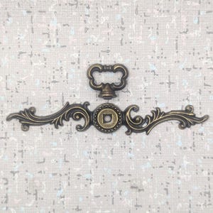 May include: An antique bronze-toned drawer pull and keyhole escutcheon set. The drawer pull has a decorative handle and a central medallion with a square design. The escutcheon has a curved, ornate design with floral accents.