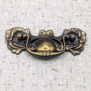 以下が含まれることがあります： 装飾的なデザインのアンティーク真鍮引き出しプル。プルは、両側に2つの華やかな花のアクセントが付いた中央のハンドルを備えています。真鍮は暗く、古くなった緑青があり、ヴィンテージな外観になっています。