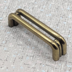 May include: A close-up of a gold-coloured metal rectangular buckle with rounded edges. The buckle has three parallel bars and is designed for use in bags, belts, or other accessories. The metal has a slightly aged appearance.